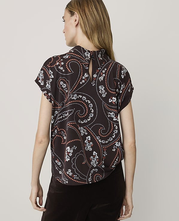 A woman in a dark brown paisley-patterned blouse stands against a plain, light background.