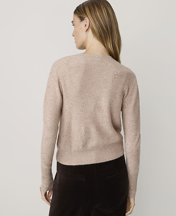 A woman in a sparkly pink cardigan and dark brown pants stands against a plain, light-colored background.