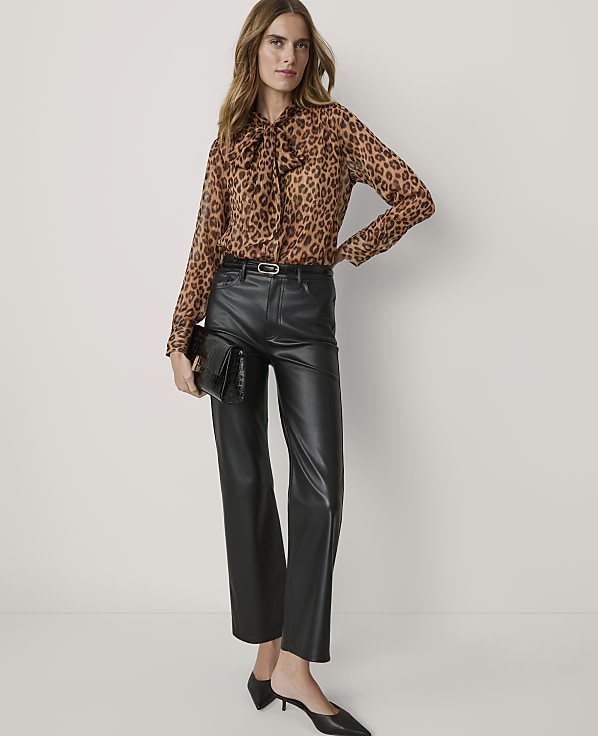 A woman in a leopard print blouse and black leather pants stands against a plain, light-colored background, holding a black clutch.
