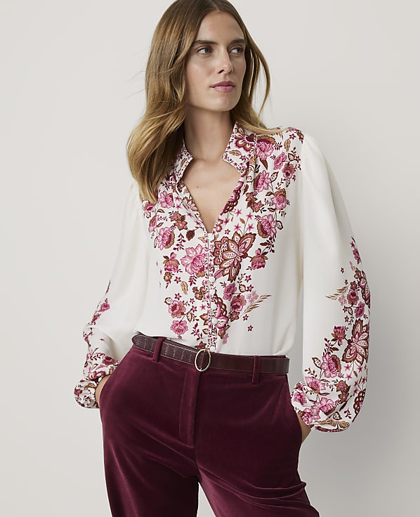 A woman in a white floral blouse and burgundy velvet pants stands against a plain background.