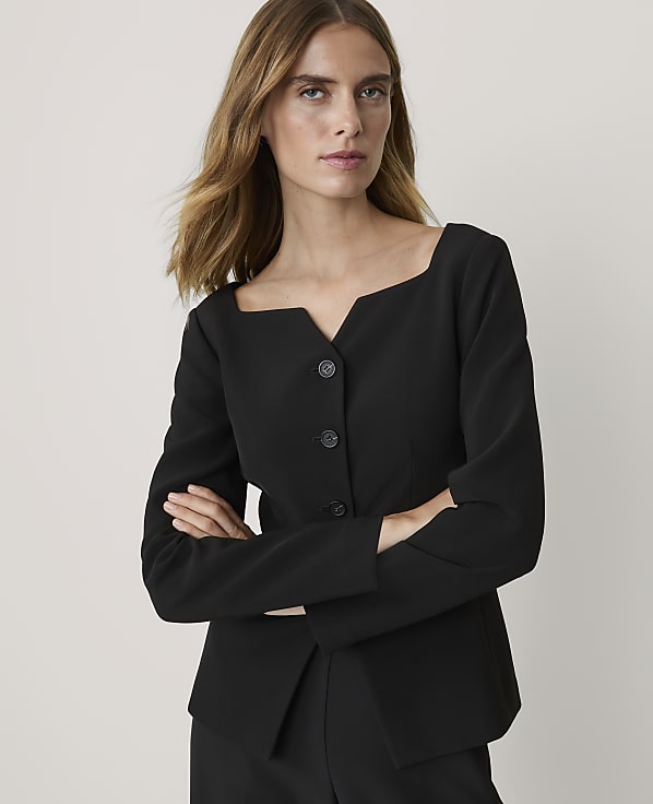 A woman in a black jacket with a square neckline stands with her arms crossed against a plain gray background.