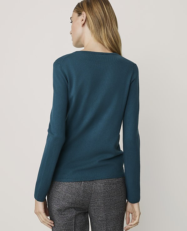 A woman in a teal sweater and patterned pants stands against a plain, light-colored background.