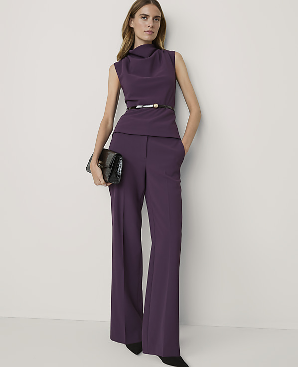A woman in a purple pantsuit with a cowl neck and a black clutch stands against a plain white background.