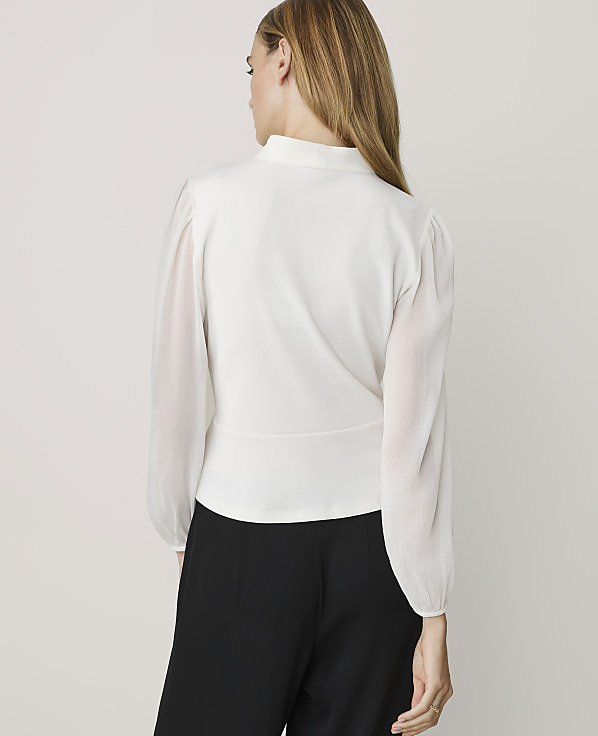 A woman in a white blouse and black pants stands against a plain white background.