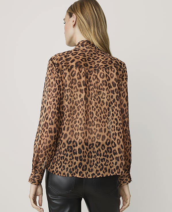 A woman in a leopard print blouse stands against a plain, light-colored background.