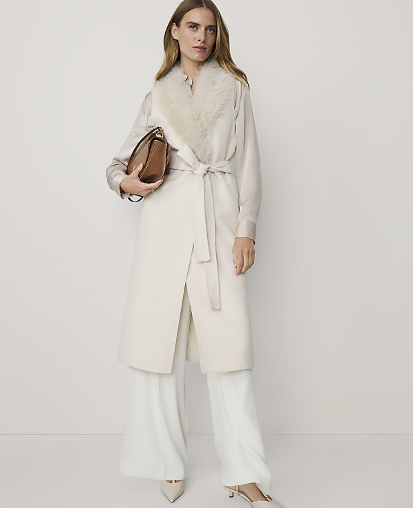 A woman in a cream coat with a fur collar and wide-leg pants stands against a plain, light background, holding a brown handbag.