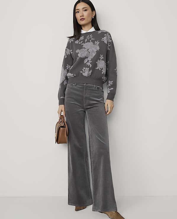 A woman in a floral sweater and gray corduroy pants stands against a plain, light-colored wall, holding a brown handbag.