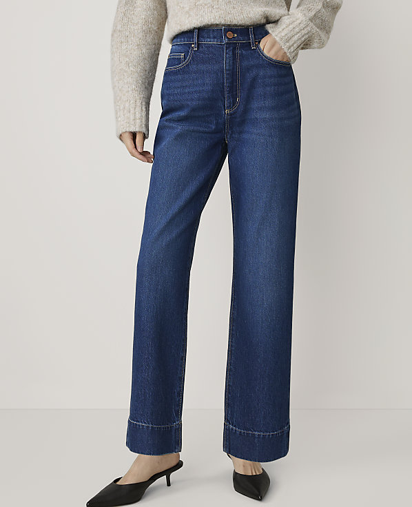 A person in a beige sweater and dark wash wide-leg jeans with cuffed hems stands against a plain white background.