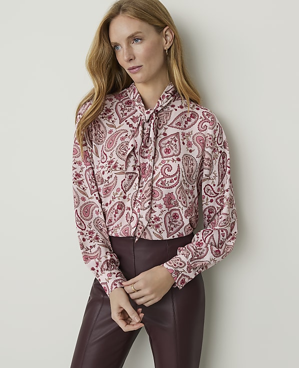 A woman in a pink paisley blouse and burgundy pants stands against a plain gray background.