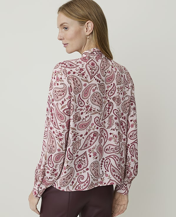 A woman in a pink paisley blouse and burgundy pants stands against a plain, light-colored background.