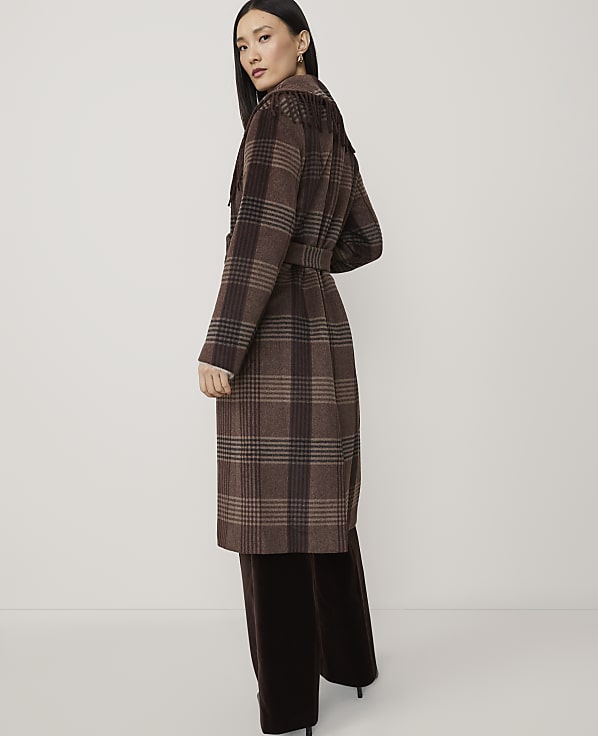 A woman in a brown plaid coat and dark pants stands against a plain, light background.