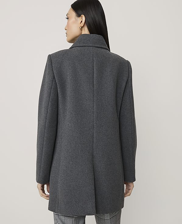 A woman in a grey coat stands against a plain grey background.