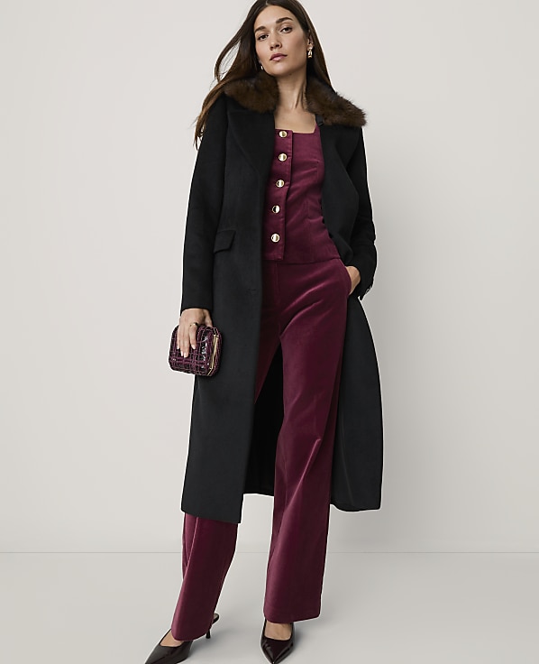 A woman in a black coat and burgundy velvet jumpsuit stands against a plain, light background, holding a small clutch.