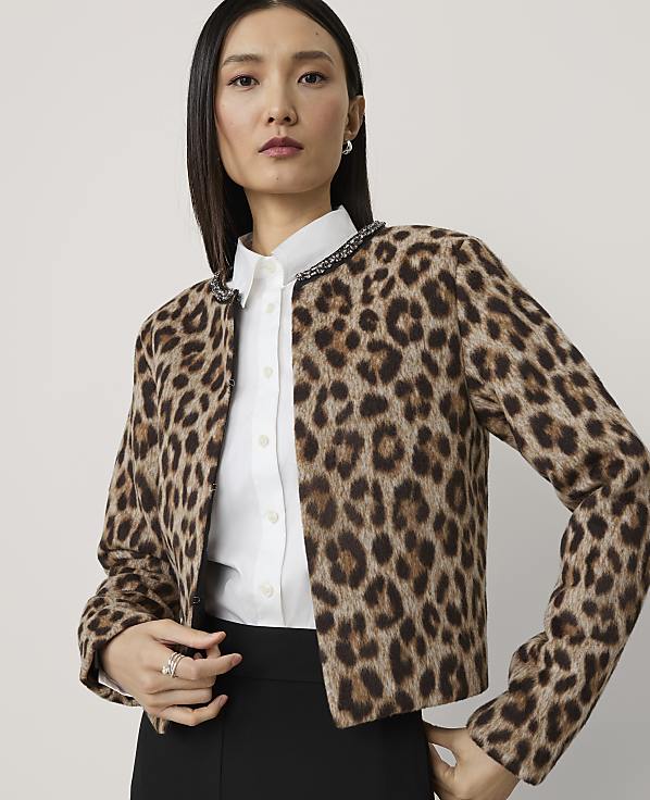 A woman in a leopard print jacket and white shirt stands against a plain, light gray background.