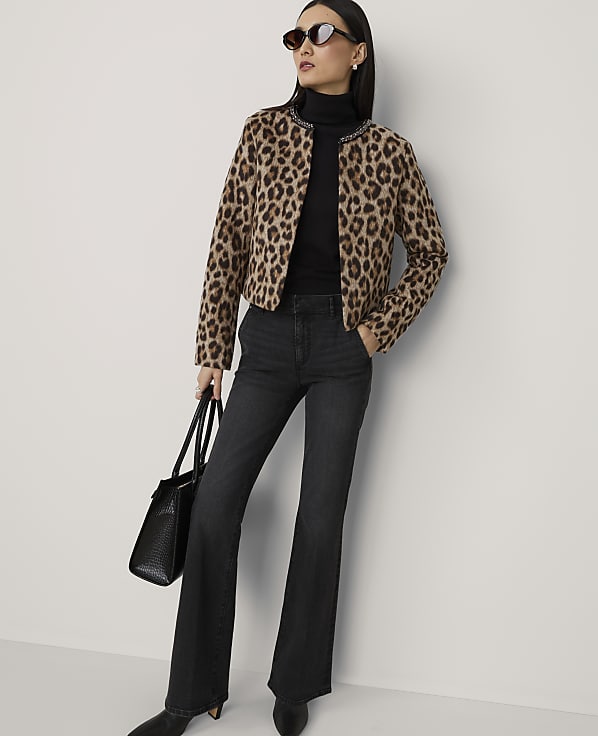 A woman in a leopard print jacket and dark flared jeans stands against a plain, light-colored wall, holding a black handbag.