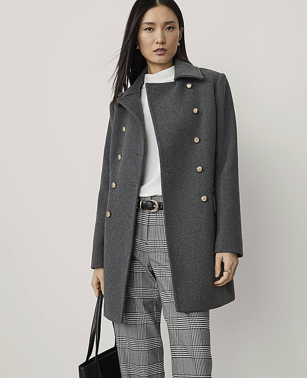 A woman in a grey double-breasted coat and plaid pants stands against a plain, light grey background, holding a black tote bag.