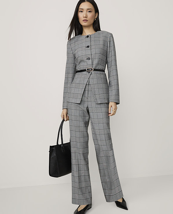 A woman in a grey plaid suit and black heels stands with a black tote bag against a plain white background.