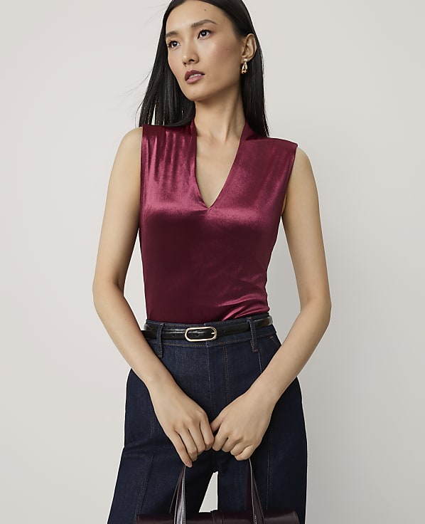 A woman in a burgundy velvet top and dark jeans stands against a plain white background, holding a dark handbag.
