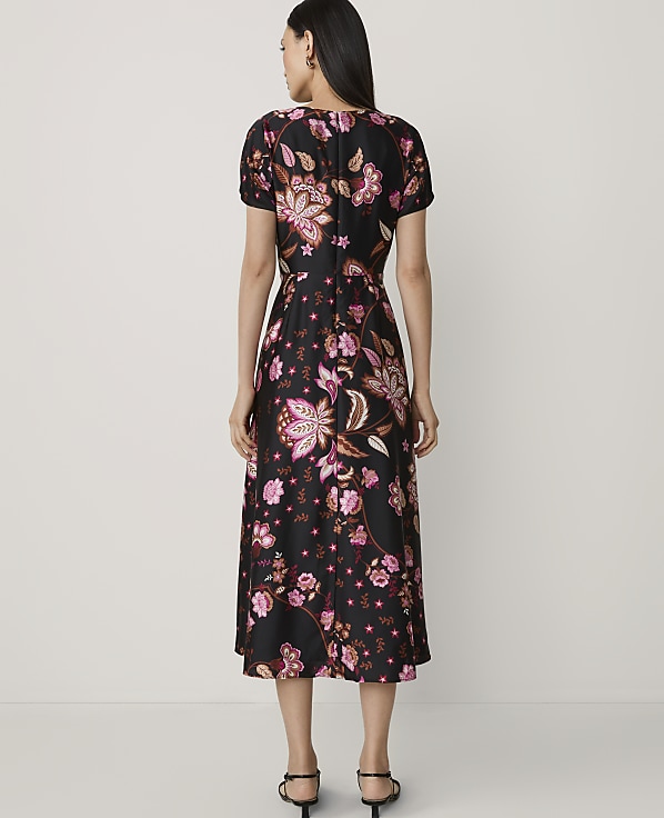 A woman in a black floral dress stands against a plain, light-colored background.