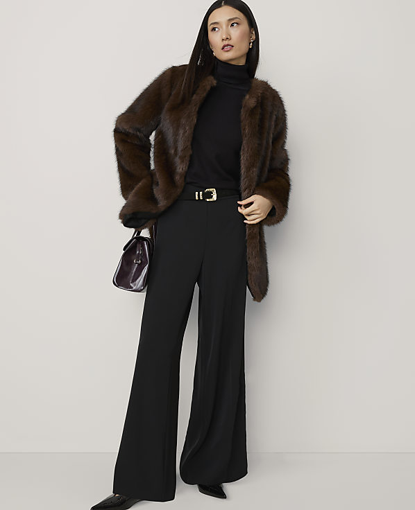 A woman in a brown fur coat, black turtleneck, and wide-leg pants stands against a plain white background, holding a dark handbag.