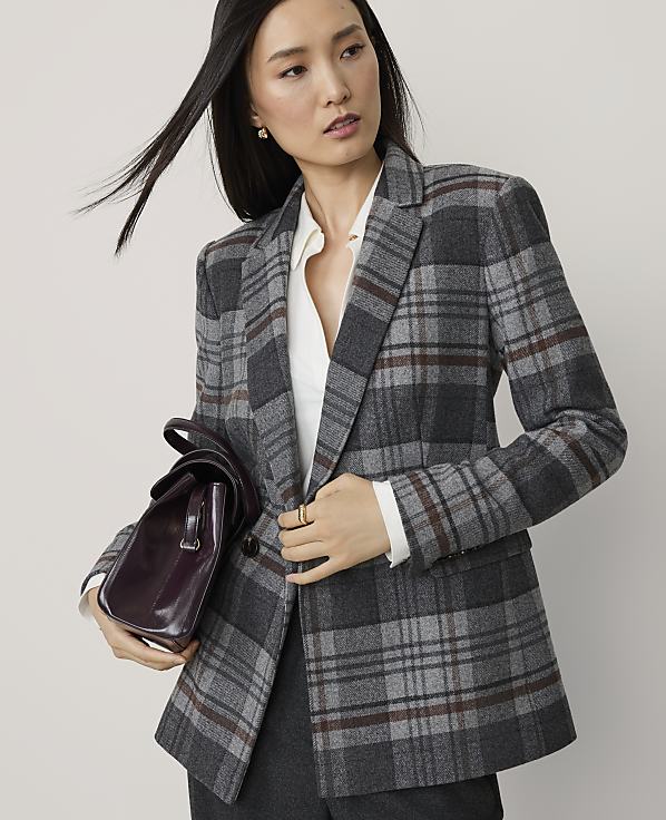 A woman in a gray plaid blazer and white blouse holds a burgundy handbag in front of a plain gray background.