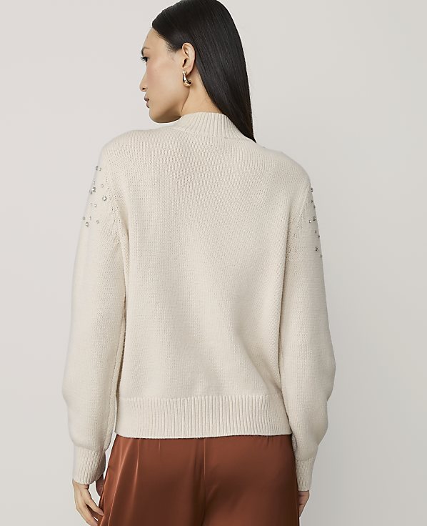 A woman in a cream-colored sweater with beaded shoulders stands against a plain white background.