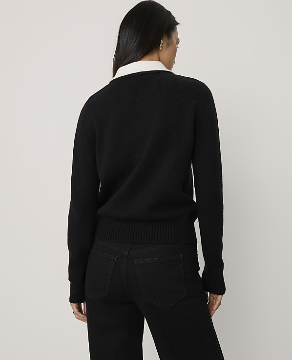A woman wearing a black sweater and black pants stands with her back to the camera against a plain, light-colored background.