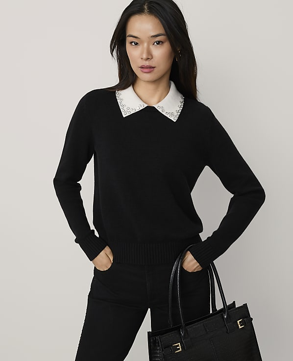 A woman wearing a black sweater with a beaded white collar and black pants holds a black handbag against a plain light gray background.