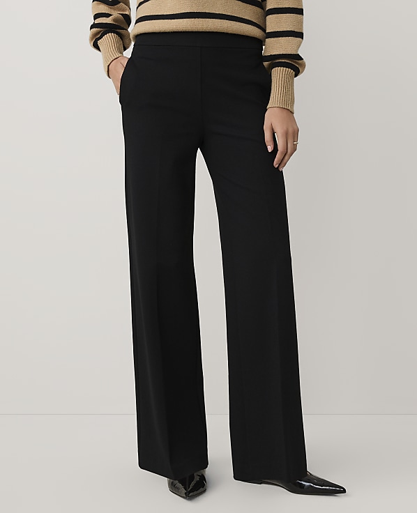 A person wearing a striped tan and black sweater with wide-leg black trousers and pointed black heels stands against a plain light gray background.