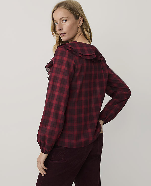 A woman wearing a red and black checkered blouse and dark burgundy trousers stands against a plain, light-colored background.