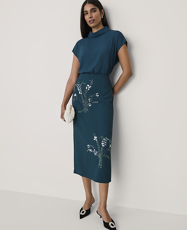 A woman wearing a teal two-piece outfit with floral sequins and black heels stands against a plain white background.