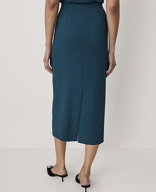 A person wearing a teal midi skirt and black pointed-toe mules stands against a plain white background.