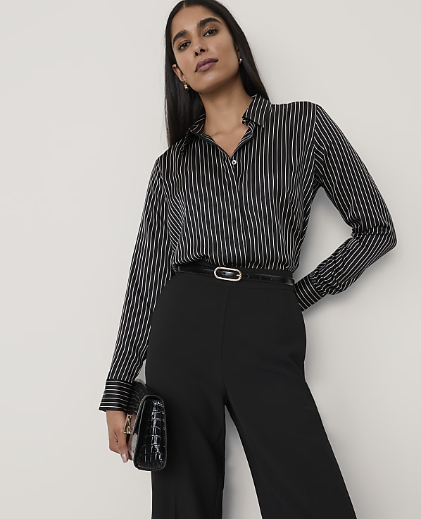 A woman wearing a black and white striped shirt and black trousers stands against a plain white background.
