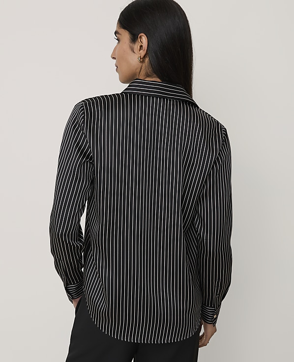A woman wearing a black and white pinstriped shirt and black trousers stands against a plain light gray background.