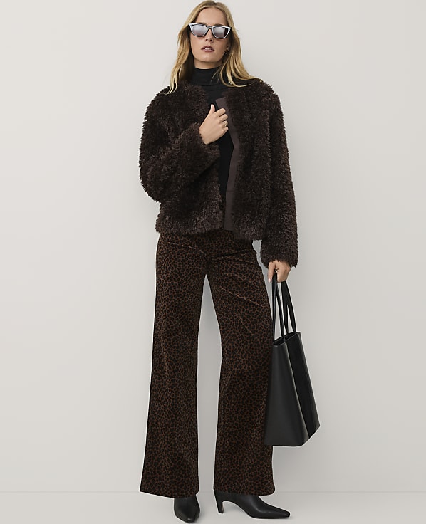 A woman wearing a brown faux-fur jacket, leopard-print trousers, and sunglasses stands against a plain white background.