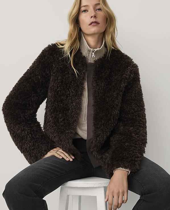 A woman wearing a dark brown faux-fur jacket and black jeans sits on a white stool against a plain light-colored background.