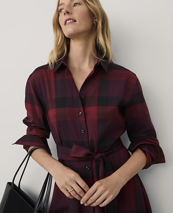 A woman wearing a red and black plaid dress holds a black handbag against a plain white background.