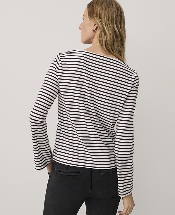 A woman wearing a black and white striped long-sleeve shirt and black jeans stands against a plain light gray background.