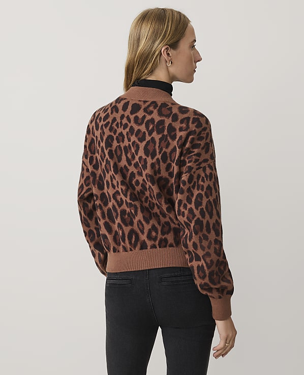 A woman wearing a leopard-print sweater and black jeans stands against a plain white background.