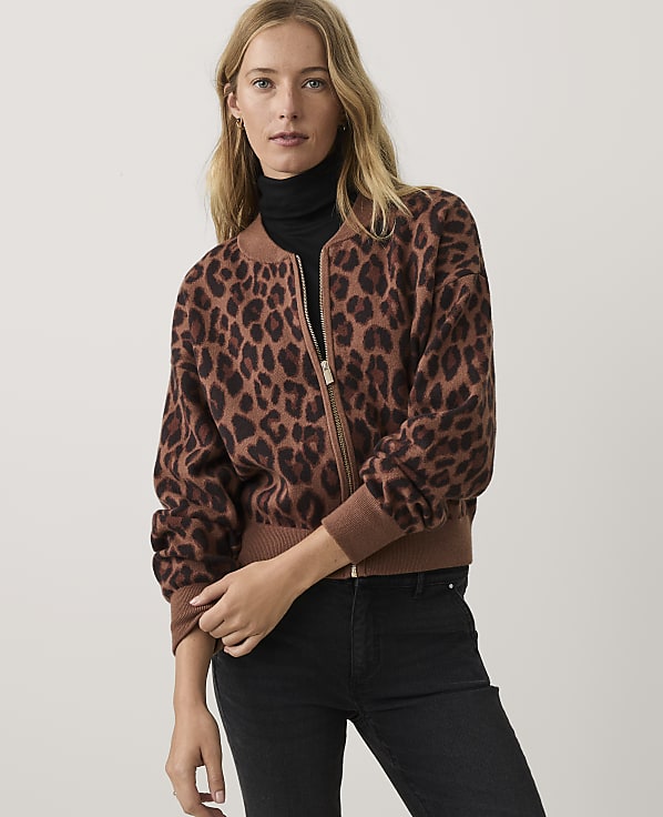 A woman wearing a brown leopard-print bomber jacket, black turtleneck, and black jeans stands against a plain light-grey background.