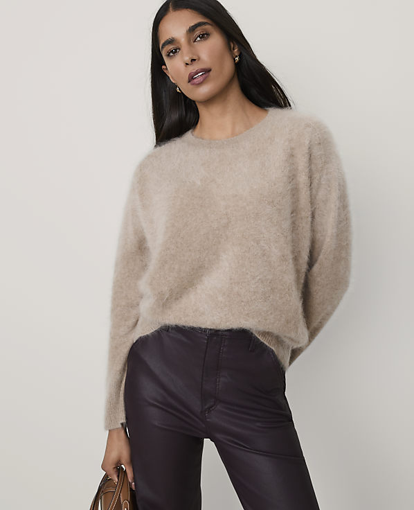 A woman wearing a fuzzy beige sweater and dark purple leather pants stands against a plain, light-colored background.