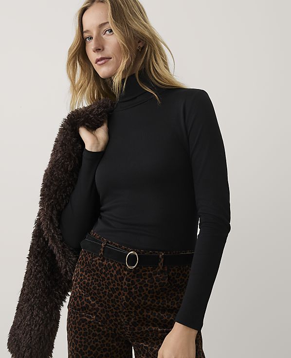 A blonde woman wearing a black turtleneck and leopard-print pants stands against a plain white background.