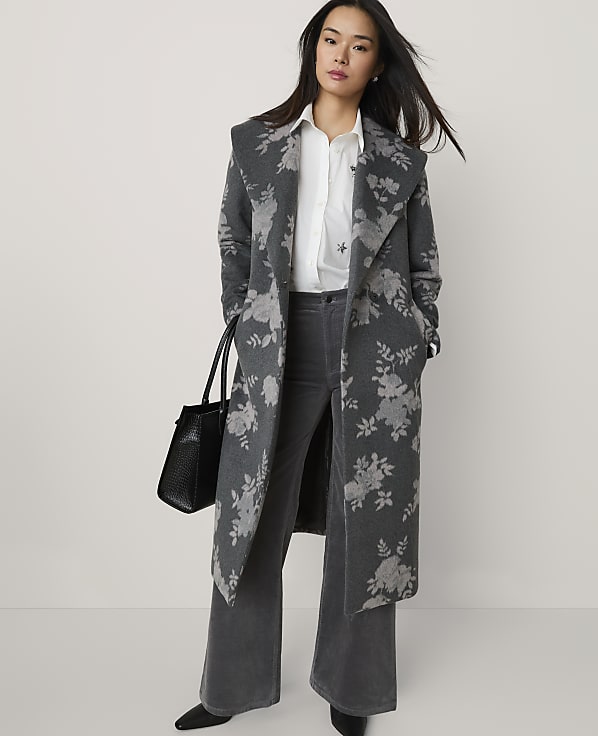 A woman wearing a grey floral coat, white shirt, and grey trousers stands against a plain light-colored background while holding a black handbag.