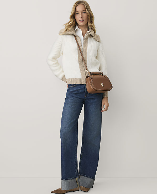 A woman wearing a white and beige zip-up sweater, blue wide-leg jeans, and a brown crossbody bag stands against a plain white background.