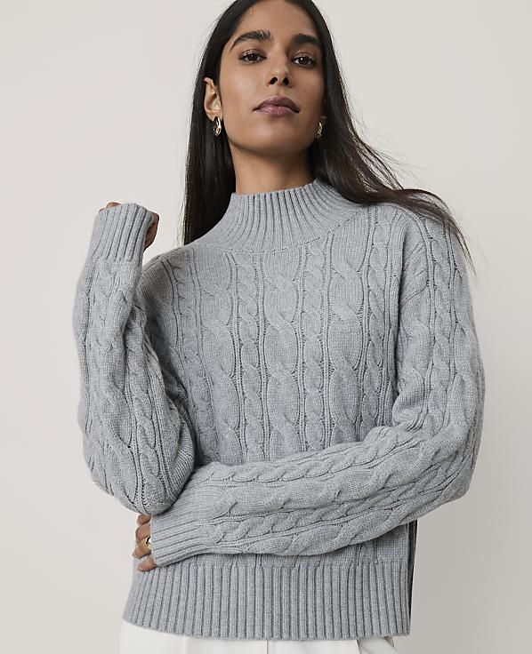 A woman wearing a grey cable-knit sweater stands against a plain, light-colored background.