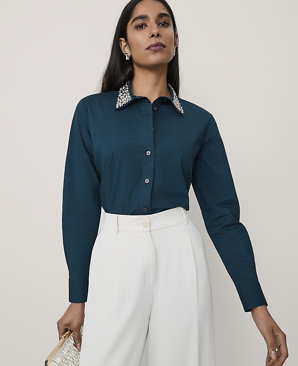 A woman wearing a teal button-down shirt with an embellished collar and white wide-leg trousers stands against a plain, light-colored background.