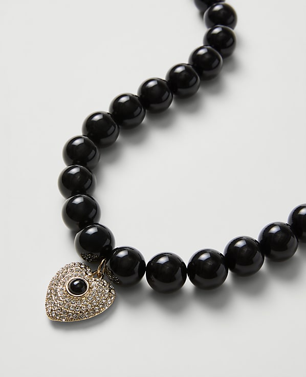 A string of polished black beads with a heart-shaped, diamond-encrusted evil eye pendant rests on a plain white background.