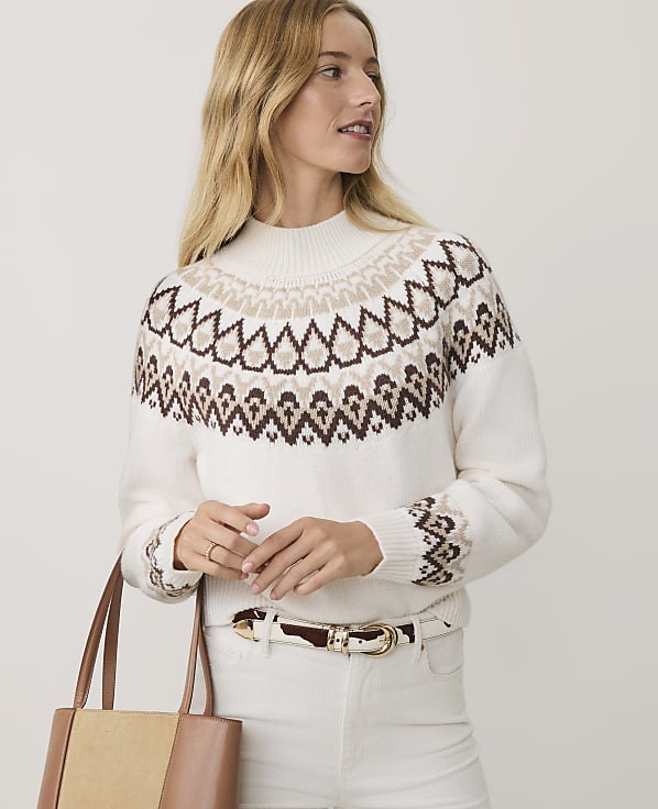 A blonde woman wearing a patterned white sweater and white pants stands against a plain light gray background.