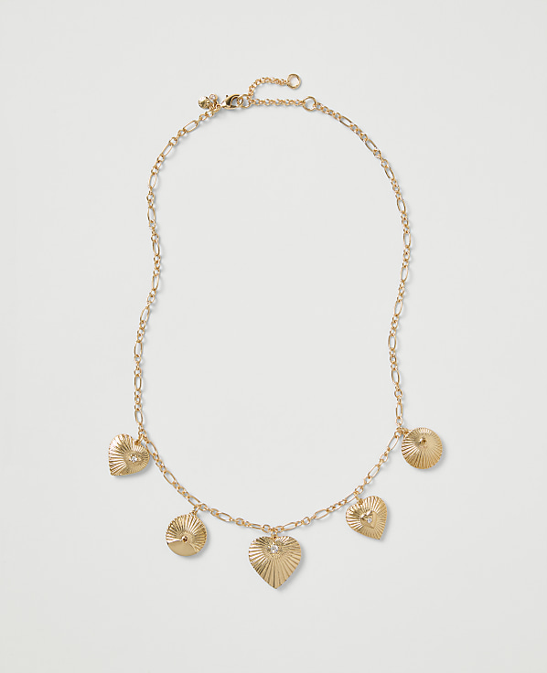 A gold chain necklace with heart and circular ribbed charms rests on a plain light gray background.