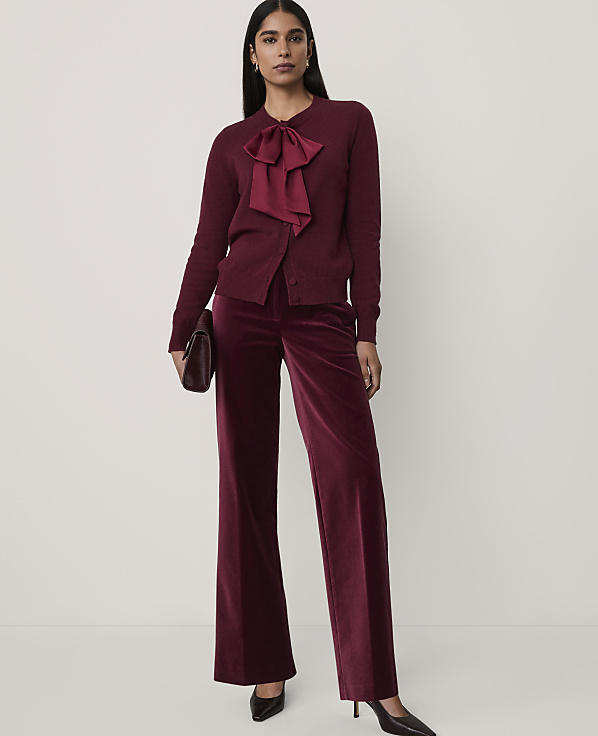 A woman wearing a burgundy bow-accented cardigan and matching velvet wide-leg trousers stands against a plain white background.